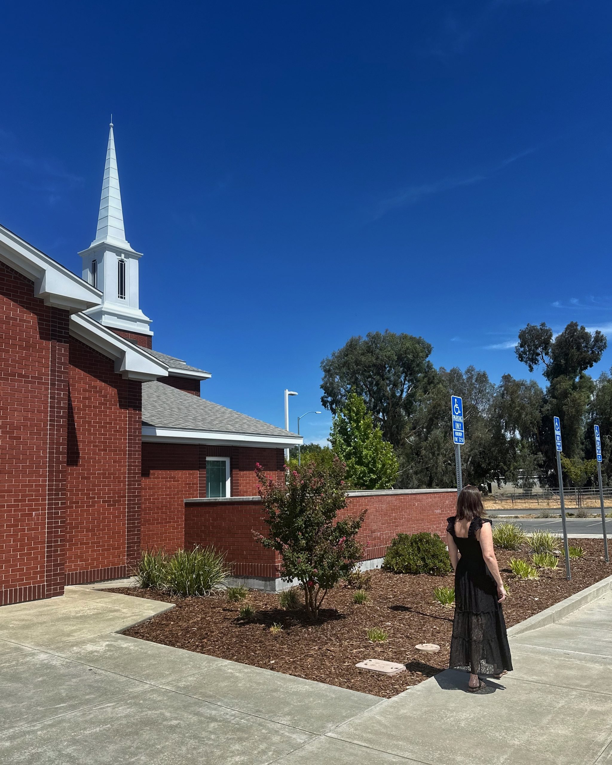 A woman in a black dress stands outside a red-brick church, looking away toward the building's steeple.